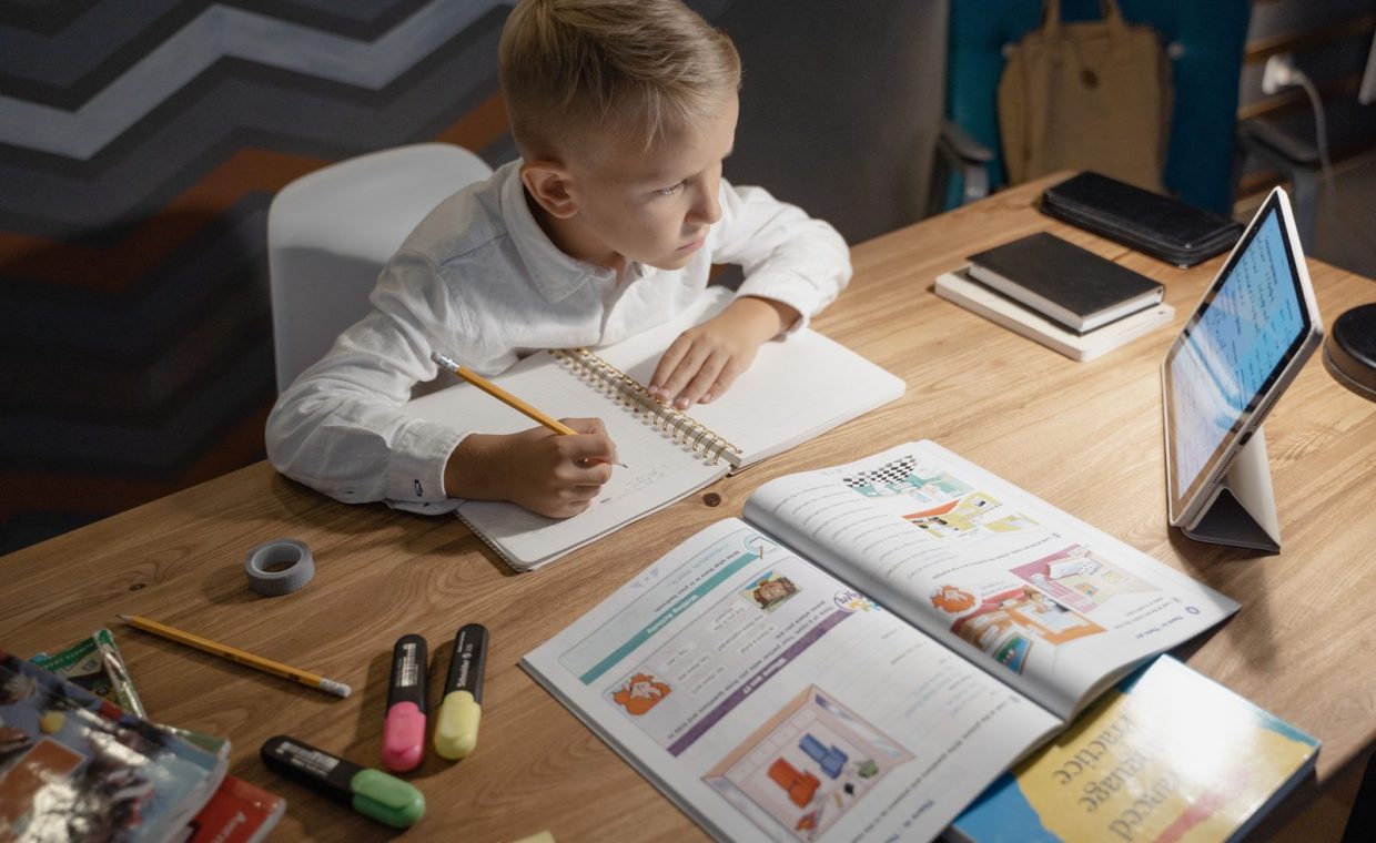 A focused young boy engages in distance learning with a tablet, books, and notebooks on a desk.