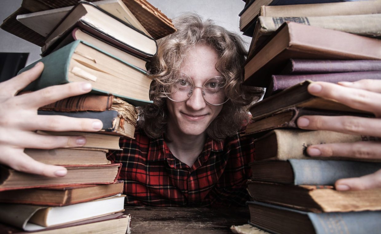 A young man in glasses surrounded by stacks of vintage books indoors, embodying knowledge and learning.
