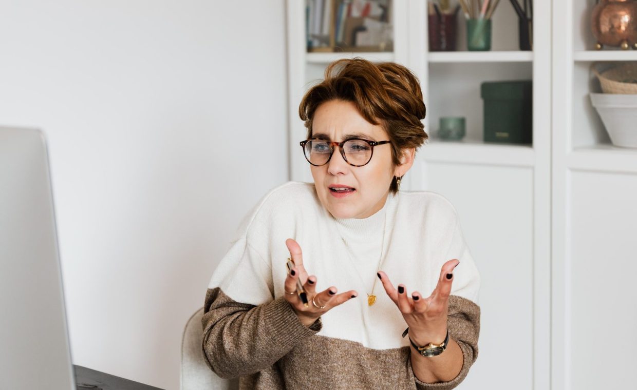 Expressive middle aged woman in casual outfit and eyeglasses gesticulating while having video conversation on computer i