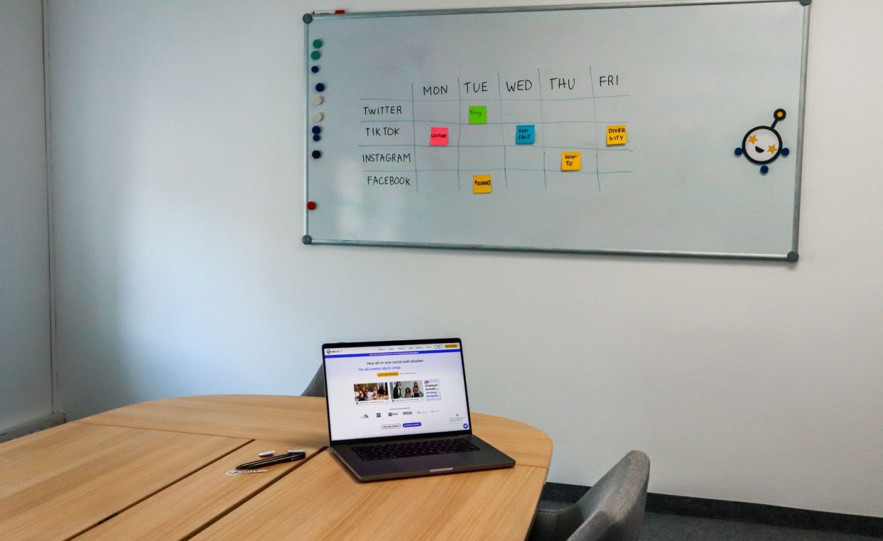 Office setup featuring a conference table, laptop, and organized whiteboard schedule, highlighting a tech-driven workspa