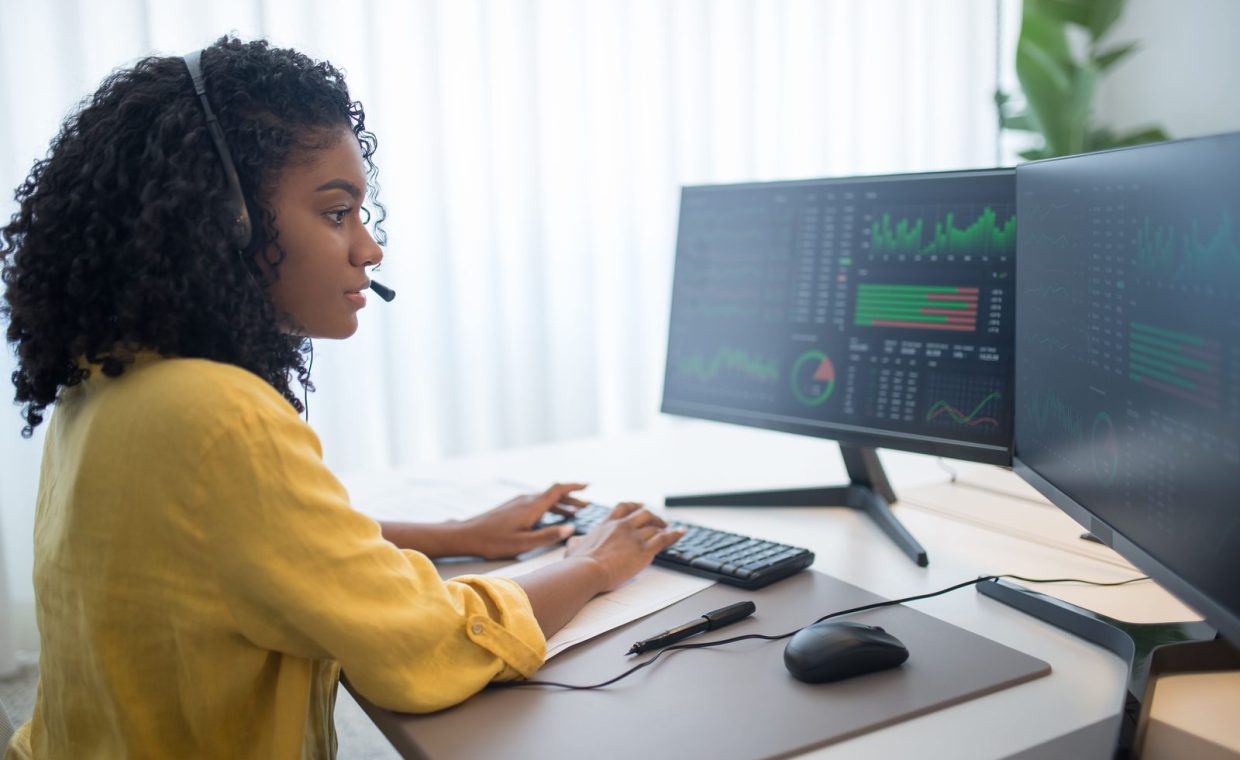 African American woman working in office analyzing data on dual computer monitors.