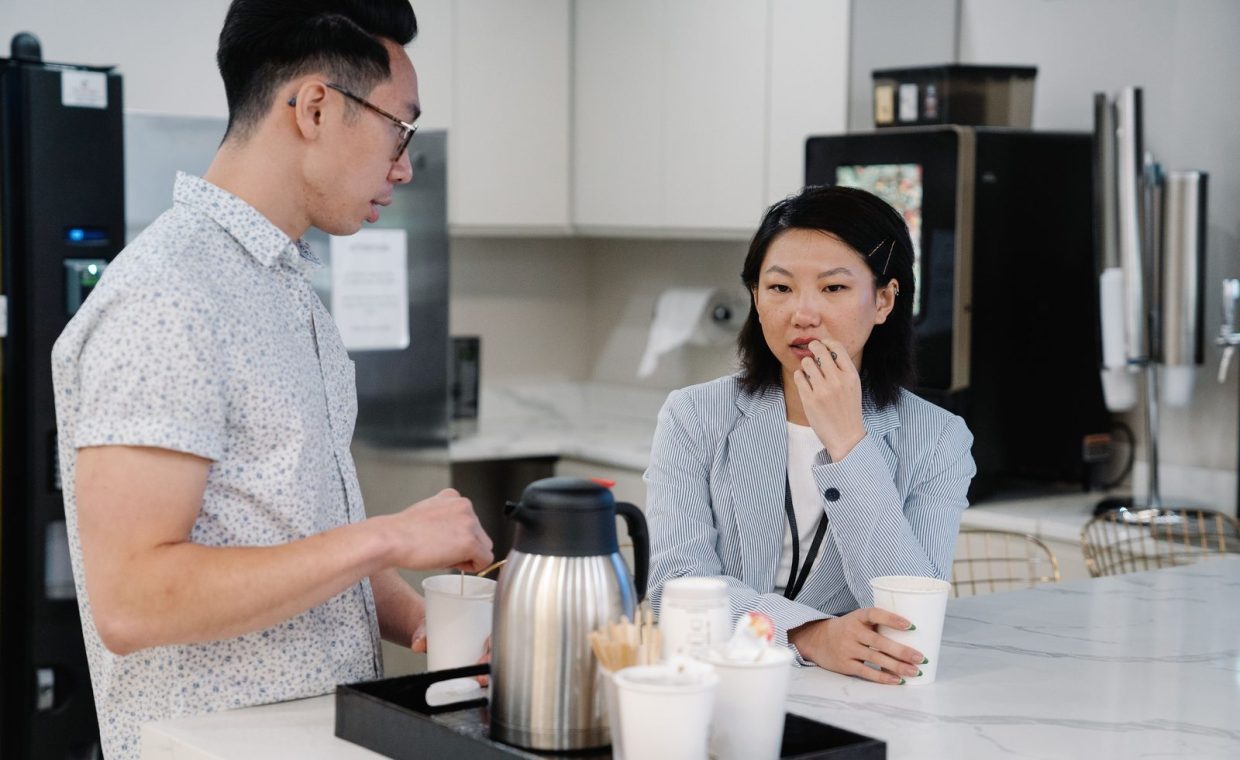 Two colleagues discussing over coffee in a modern office break room setting.