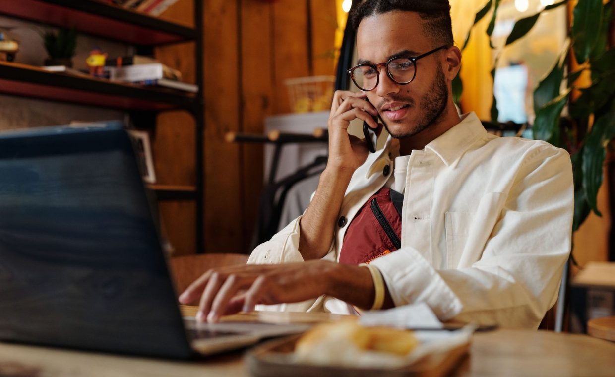 Stylish young man multitasking on laptop and phone in a cozy café interior.