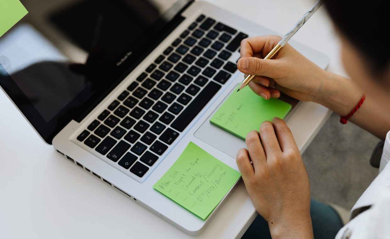 An individual writes notes on green sticky notes placed on a laptop in a work setting.