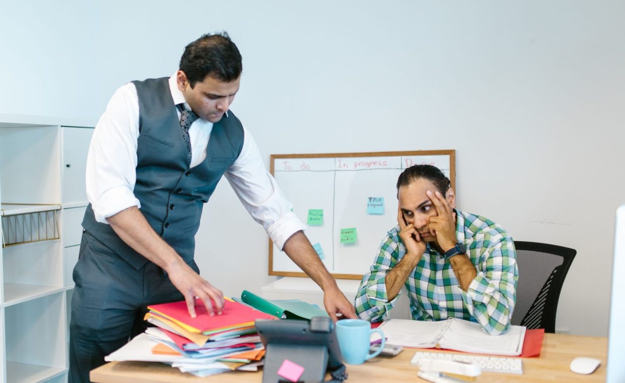 Two businessmen in an office, one stressed at the desk while another points at folders, discussing workload.