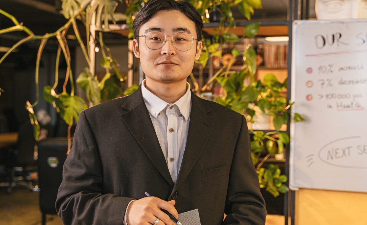 A confident young businessman in a suit and eyeglasses standing in a modern office.