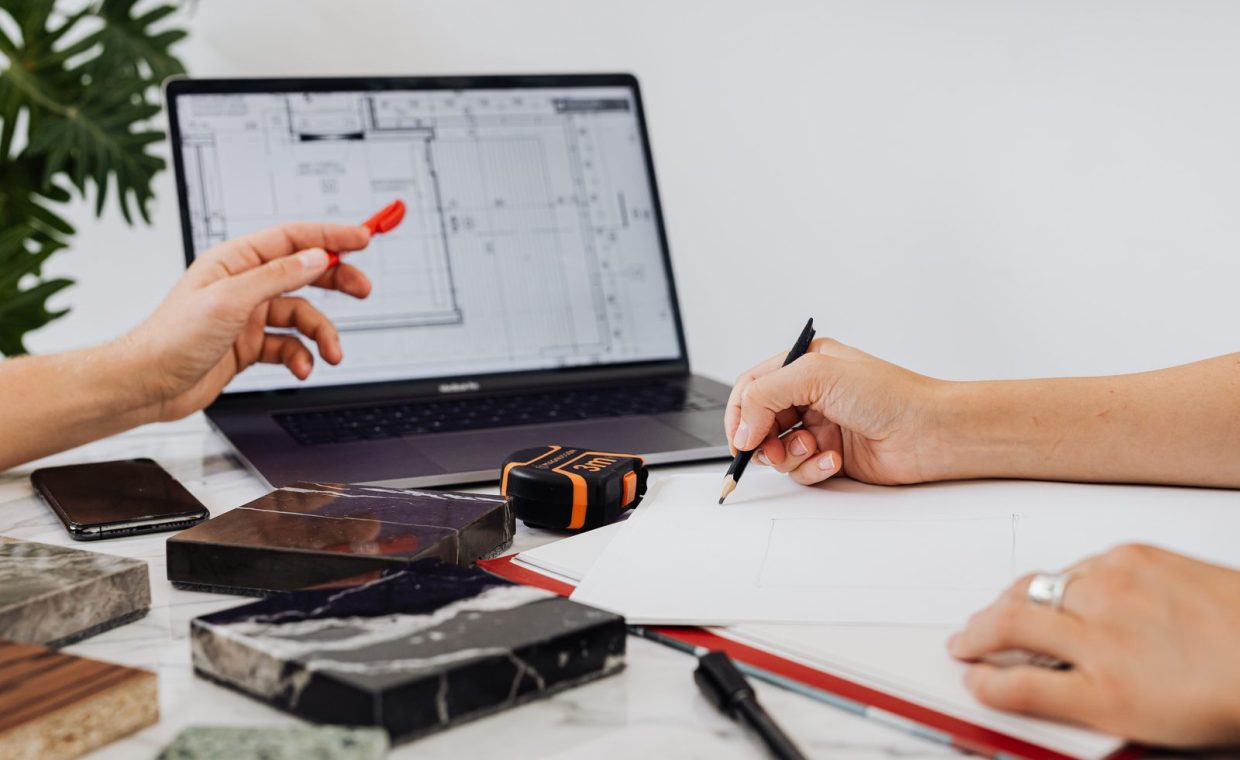 Close-up of hands sketching design plans with tile samples and a laptop, ideal for interior design contexts.
