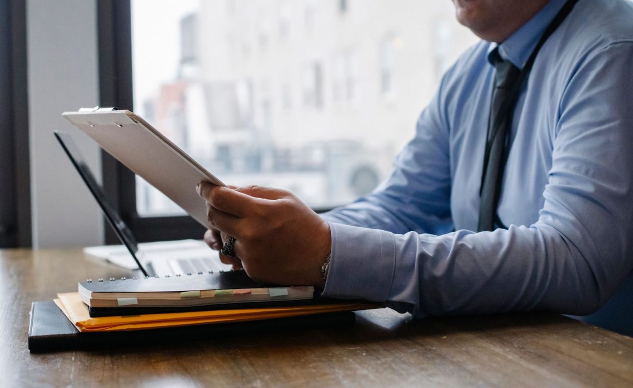 Crop unrecognizable male manager wearing blue shirt and black tie sitting at wooden desk and reading financial informati