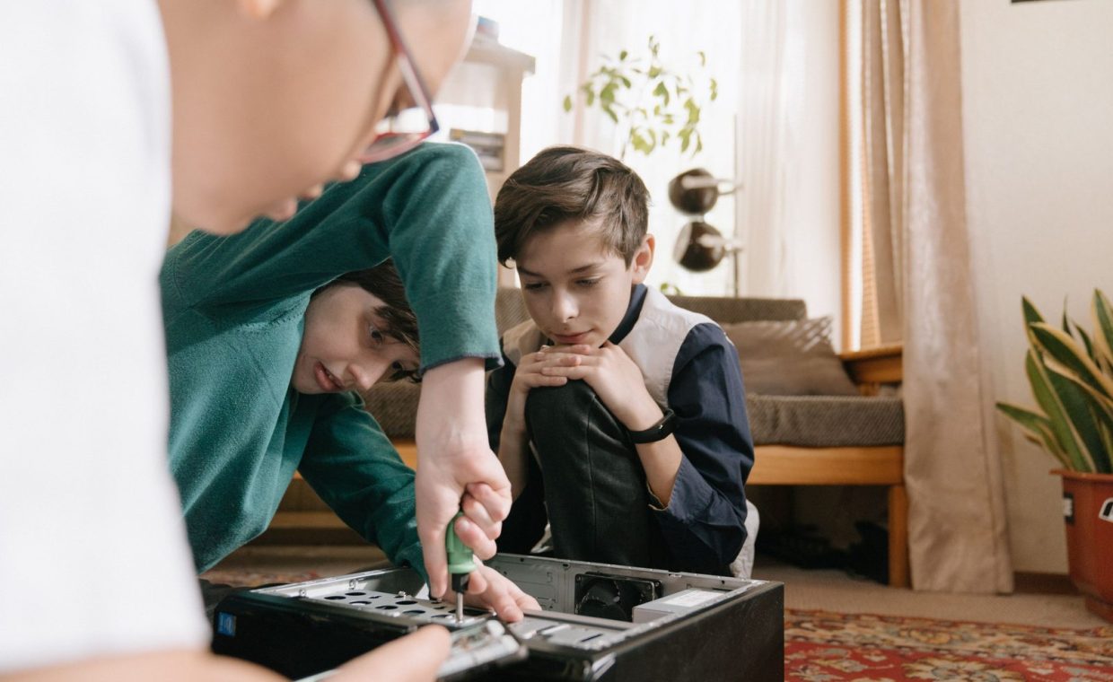 Three children collaborating to repair a computer, using tools indoors.