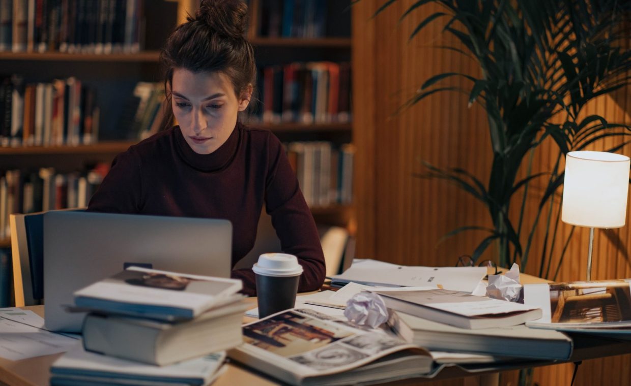 Woman working late at a desk filled with books, laptop, and coffee in a cozy library setting.