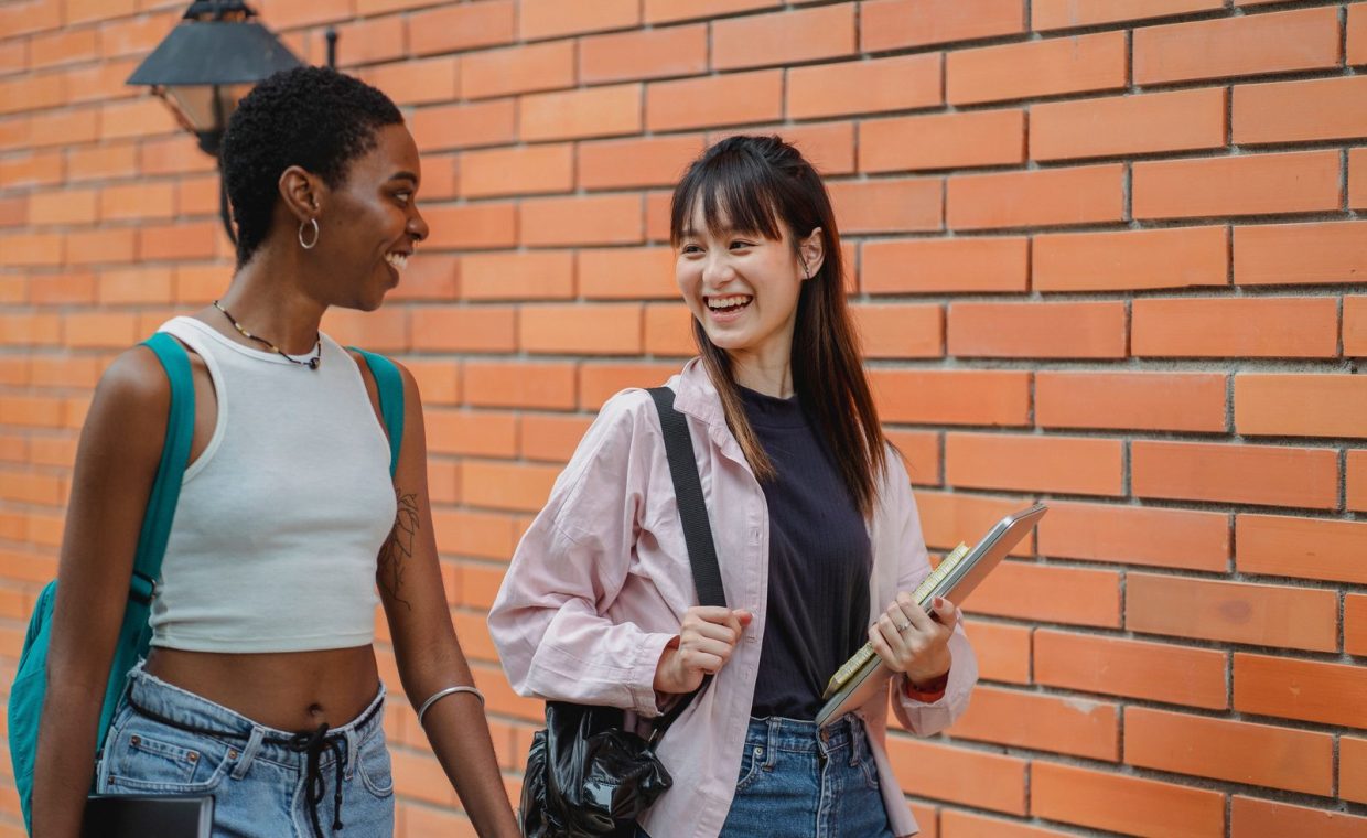 Cheerful multiracial female students wearing casual clothes carrying backpack and notebooks near college brick wall whil