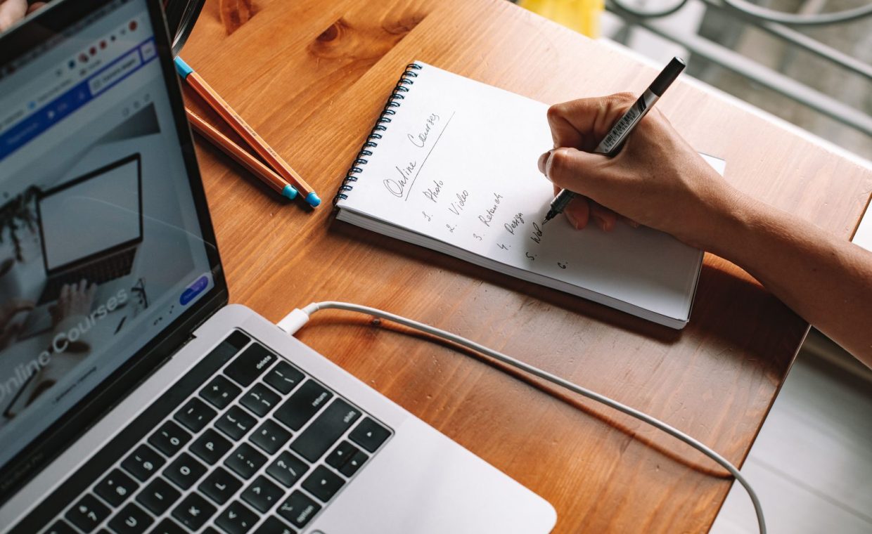 A person writing notes while researching online courses on a laptop in a home setting.