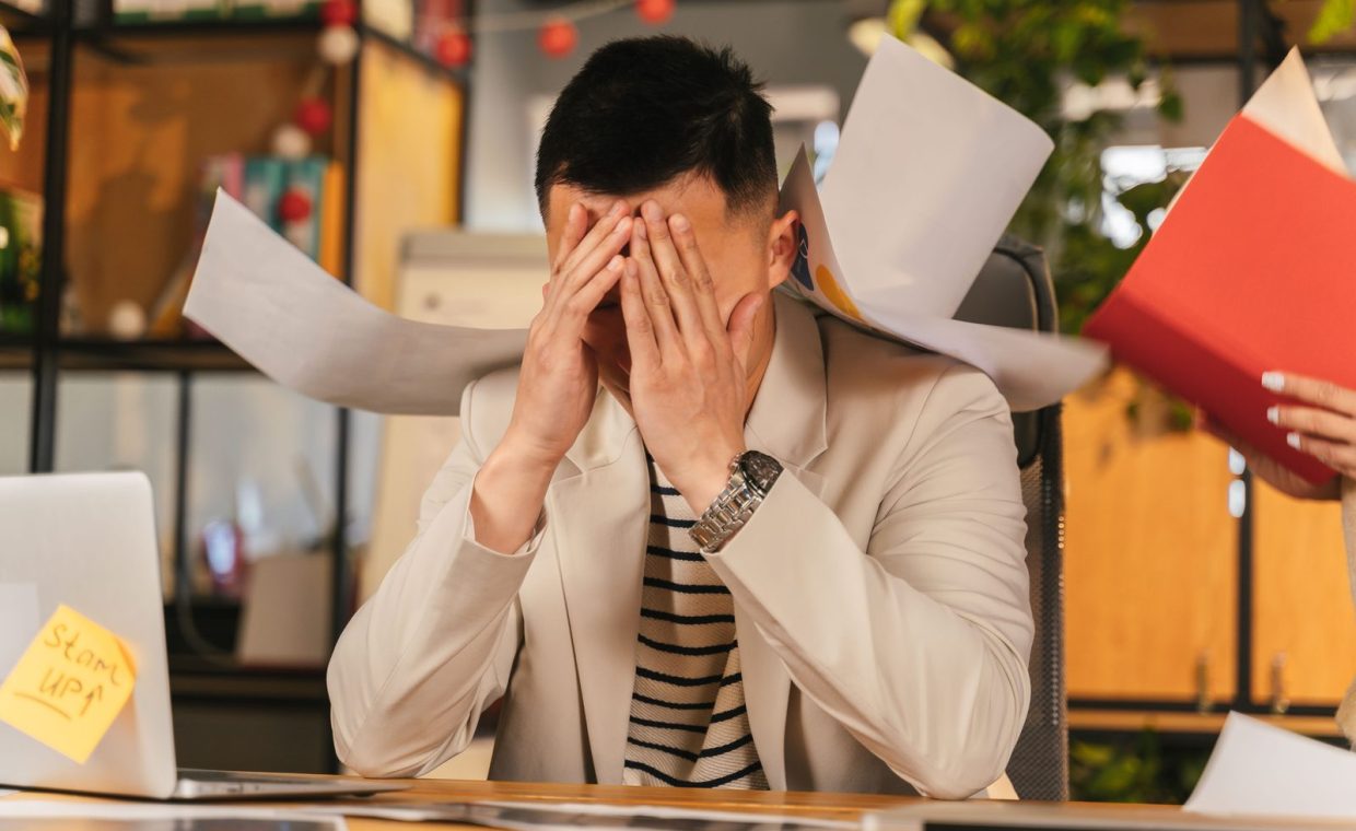 Stressed businessman overwhelmed by paperwork in office environment, demonstrating burnout.