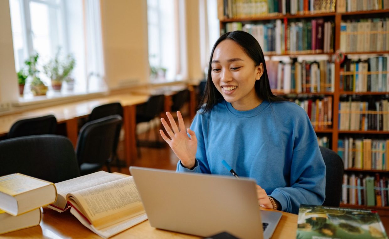 Smiling student using laptop in a library for studying and video calls.