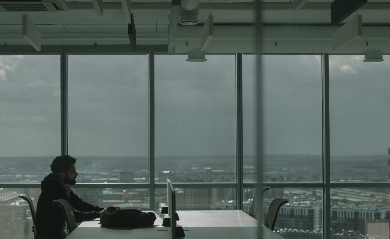 Man in modern office setting at a desk overlooking a cityscape through large windows.