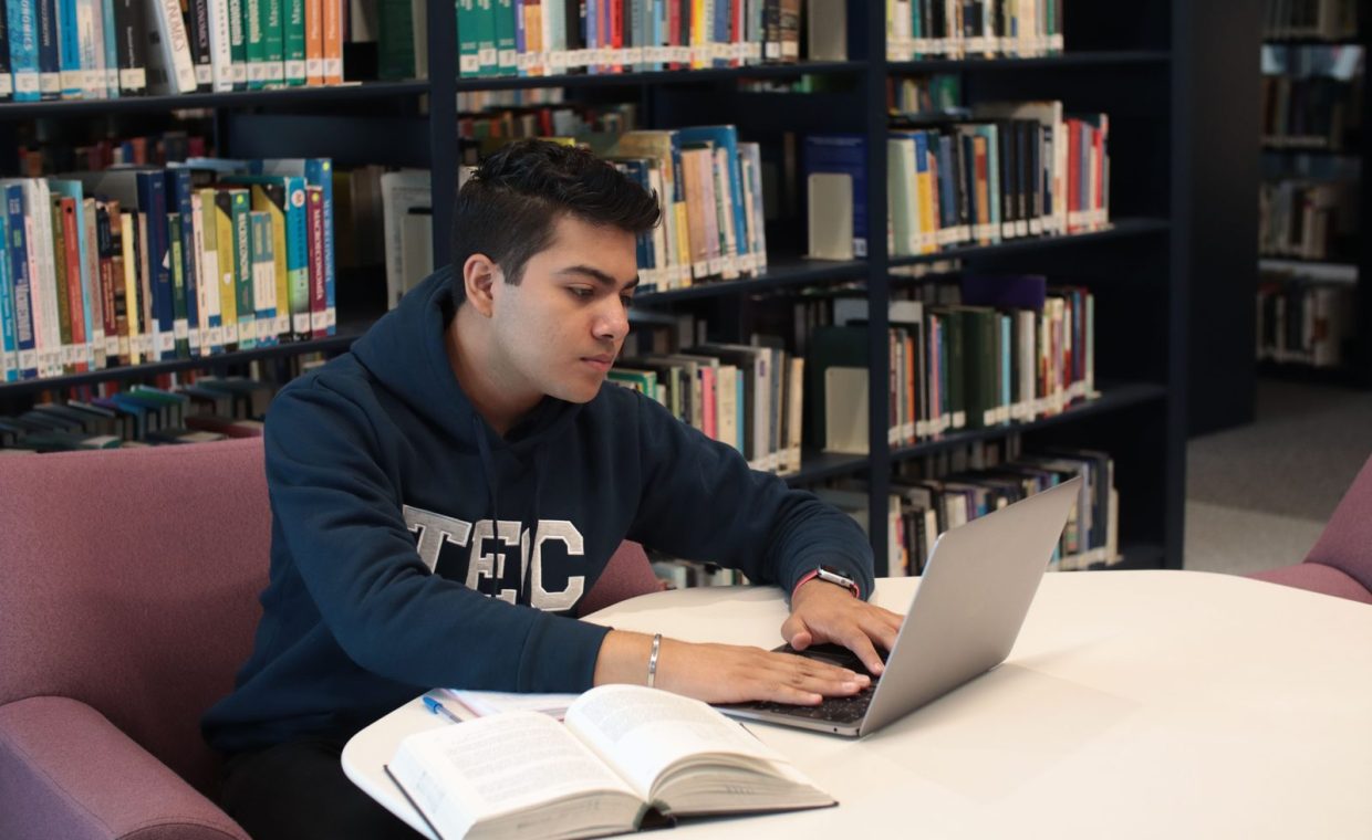 Student in library using laptop and textbooks for study and research.