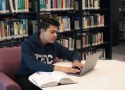 Student in library using laptop and textbooks for study and research.